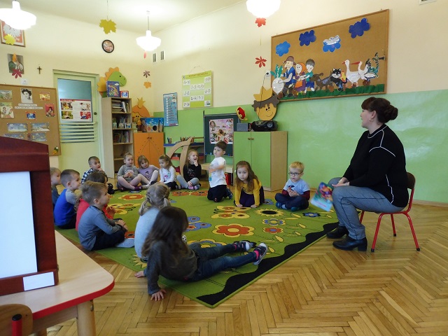 Dzieci w sali przedszkolnej siedzące na dywanie, słuchające prowadzącej nauczycielki