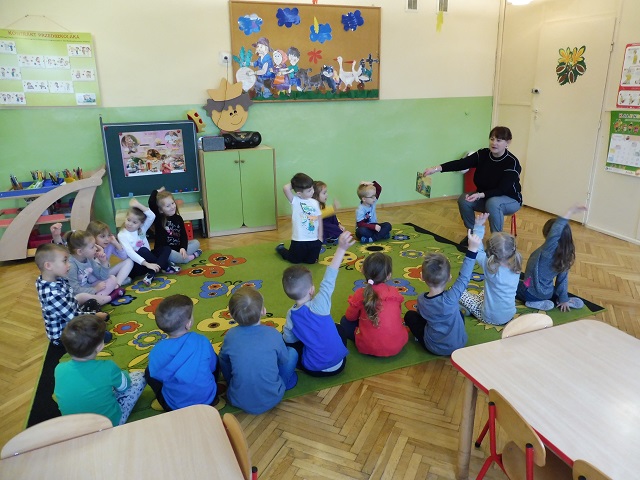 Dzieci w sali przedszkolnej siedzące na dywanie, słuchające prowadzącej nauczycielki