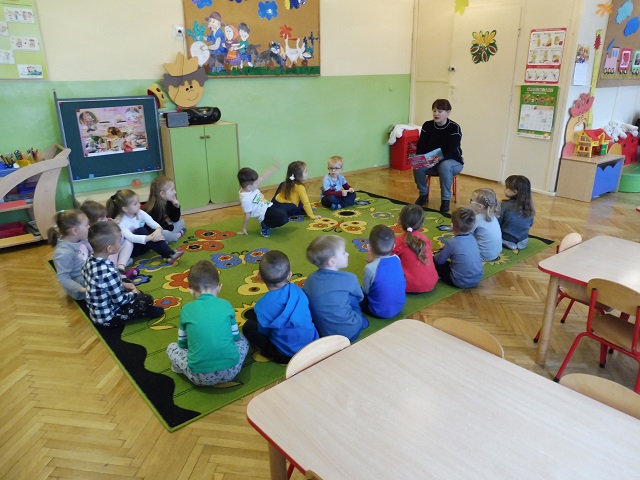 Dzieci w sali przedszkolnej siedzące na dywanie, słuchające prowadzącej nauczycielki